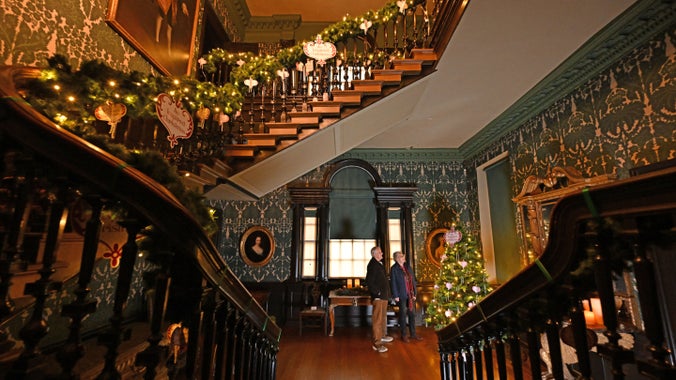 View across a banisters with garlands on, to two people looking at a decorated Christmas tree in an opulent hallway